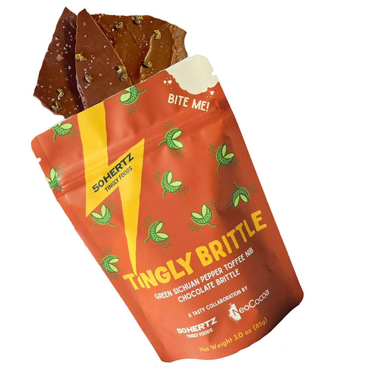 Brittle with green peppercorns spilling out of standup pouch packaging. Pouch is co branded with 50Hertz and NoCocoa with brown background and yellow and white text "Tingly Brittle Green Sichuan Paper Tofee Nib Chocolate Brittle" 