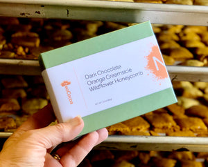 2 dimensional angle of closed box with title of "Dark Chocolate Orange Creamsicle Wildflower Honeycomb" and NeoCocoa logo. Piles of naked honeycomb candy on sheet pans in the background.