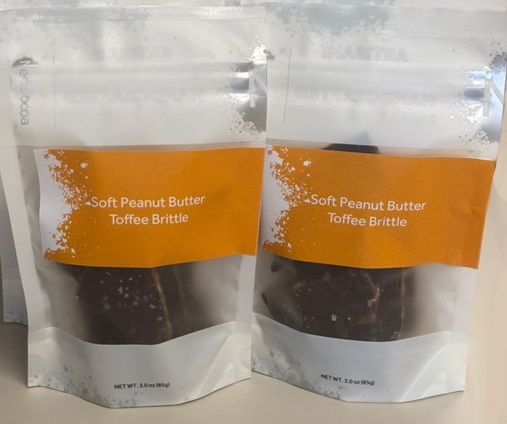 Two stand-up pouches of soft peanut butter toffee brittle cookies on a light beige background. The packaging is white with a prominent orange label containing white text that reads 'Soft Peanut Butter Toffee Brittle'.