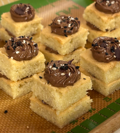 Close-up of several square sponge cake pieces topped with piped chocolate frosting and sprinkled with black and white sesame seeds, arranged on a silicone baking mat.