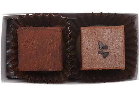 Above view and inside the rectangle box, 1 cube shaped dark salted caramel truffle (coated in cocoa powder) and 1 cube shaped milk caramel truffle with a few flakes of black salt in the center. Each truffle is in a brown paper candy cup. 