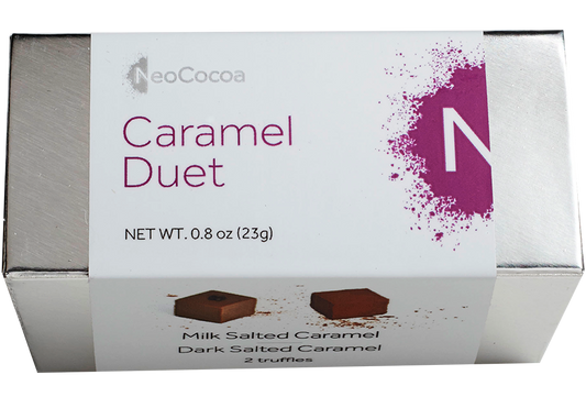 2 dimensional silver rectangle box with a label wrapped around the box stating, “Caramel Duet” with NeoCocoa logo and net weight  0.8 oz (23g). Can see a little of the side of the label with image of one dark salted caramel truffle and one milk salted caramel truffle.  